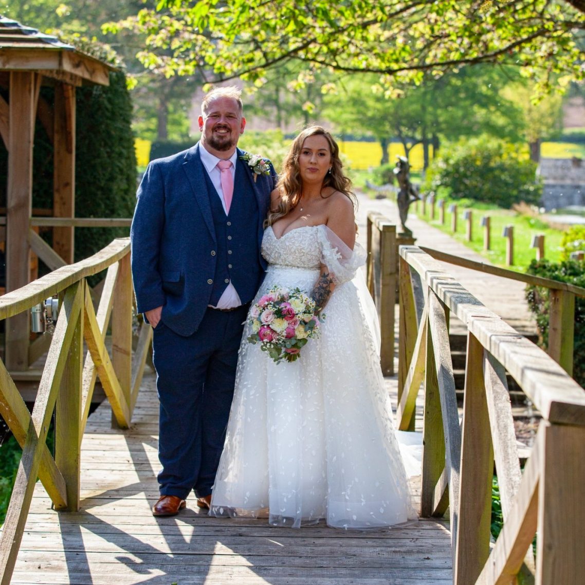 Wedding couple portrait on a bridge A bride and groom stand on a wooden bridge, surrounded by greenery and blooming trees.