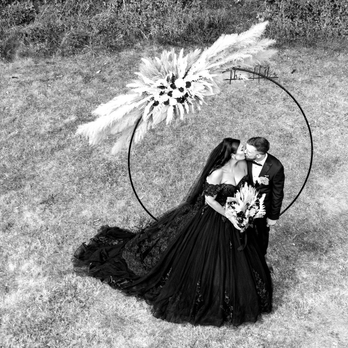 Black wedding dress A wedding couple share a kiss under a floral arch in a grassy setting.