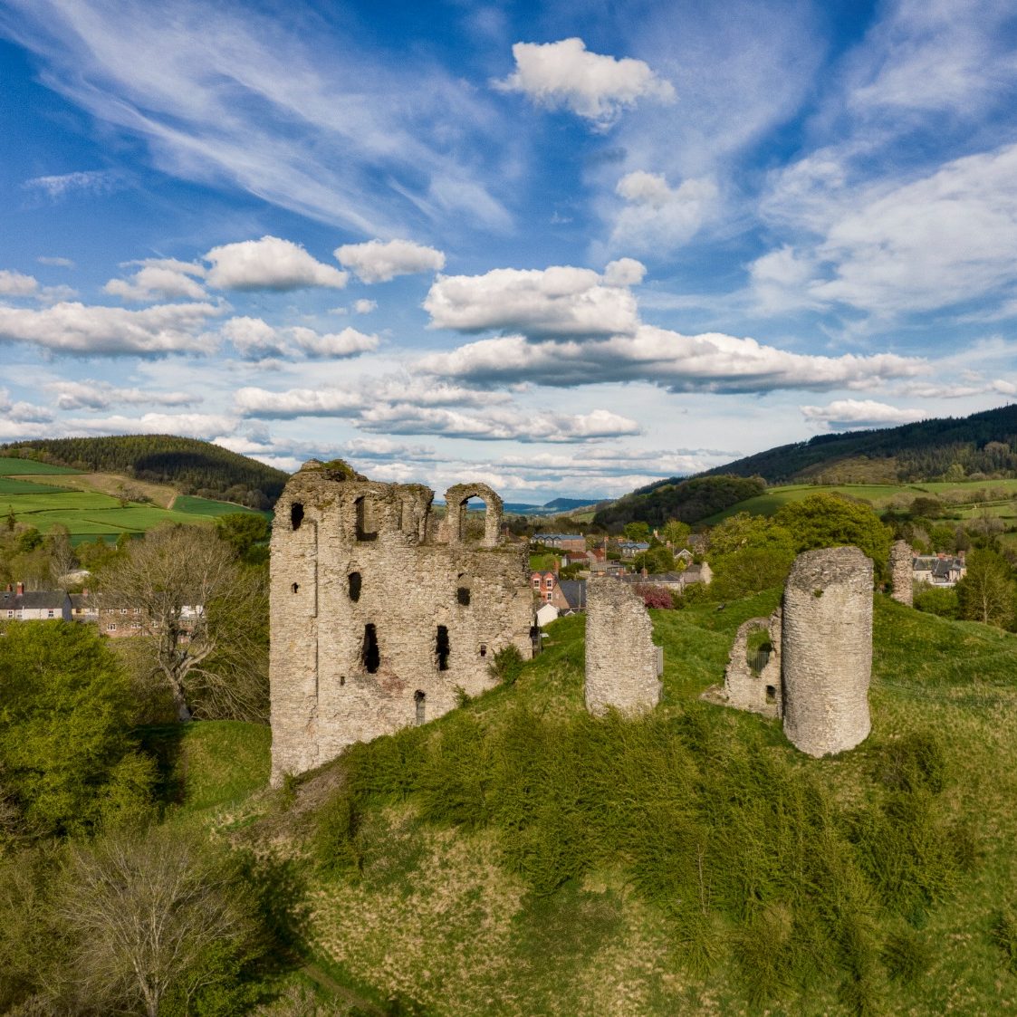 Clun Castle Ruined castle on a hill in Shropshire surrounded by greenery and hills under a cloudy sky.